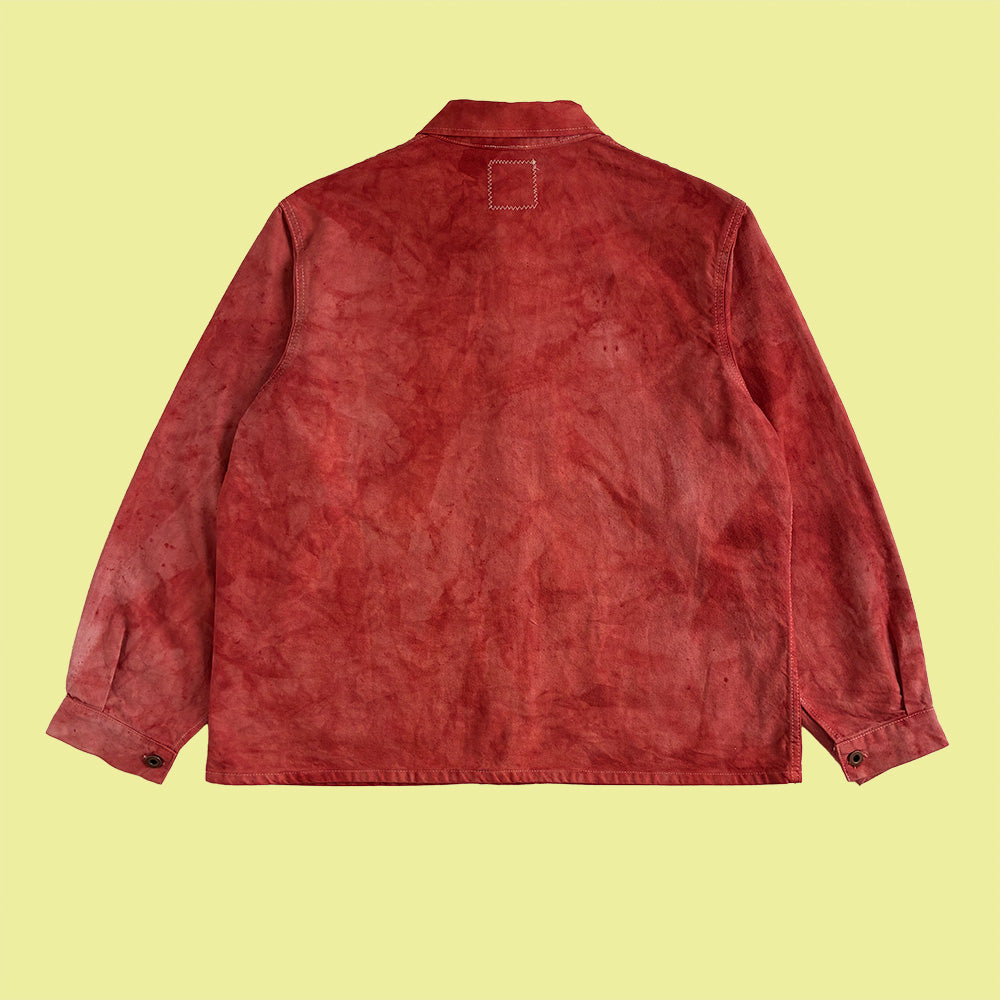 A hand dyed red and pick washed cotton chore coat shown from the back, laid flat against a pale yellow background.