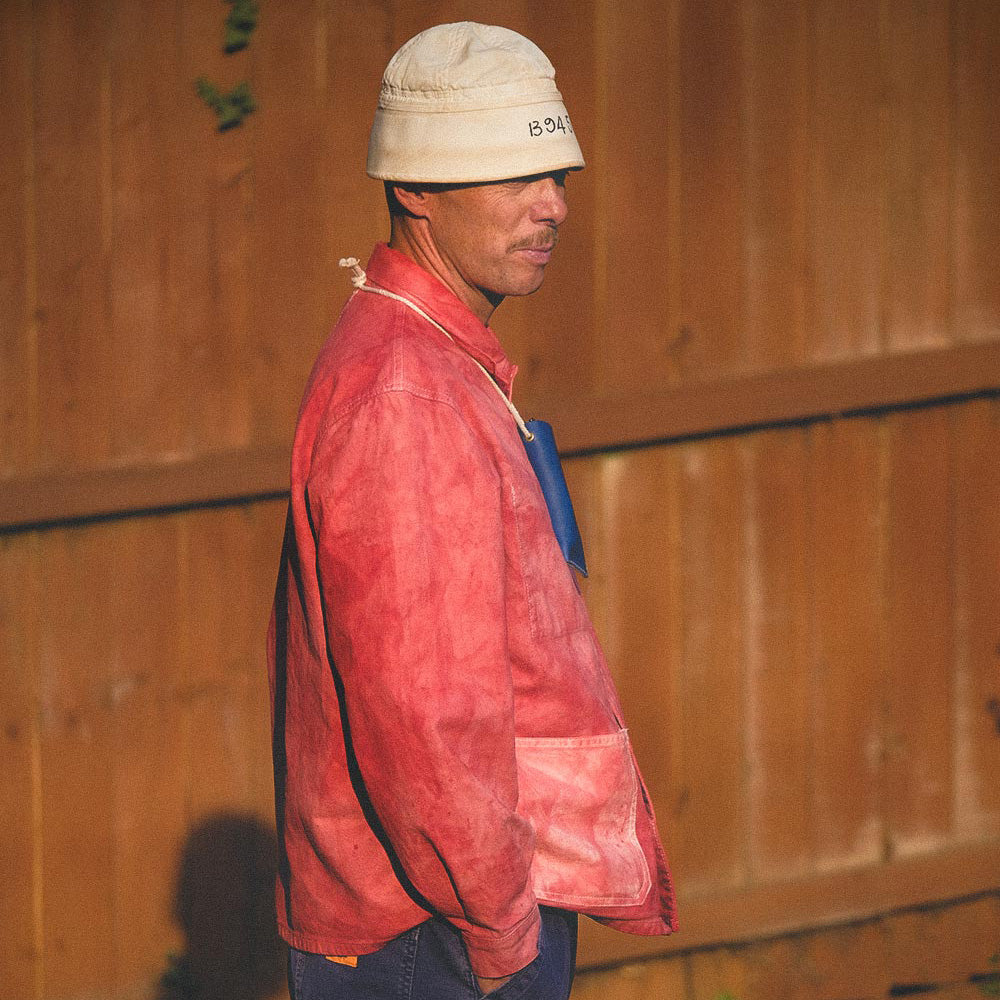 A man in profile wearing a beige bucket hat, a hand dyed chore coat, and a blue shirt, standing in front of a wooden fence.