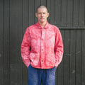 A man wearing a pink and red hand dyed chore jacket with blue jeans, standing in front of a dark gray wooden plank wall.