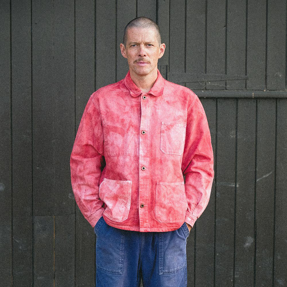 A man wearing a pink and red hand dyed chore jacket with blue jeans, standing in front of a dark gray wooden plank wall.