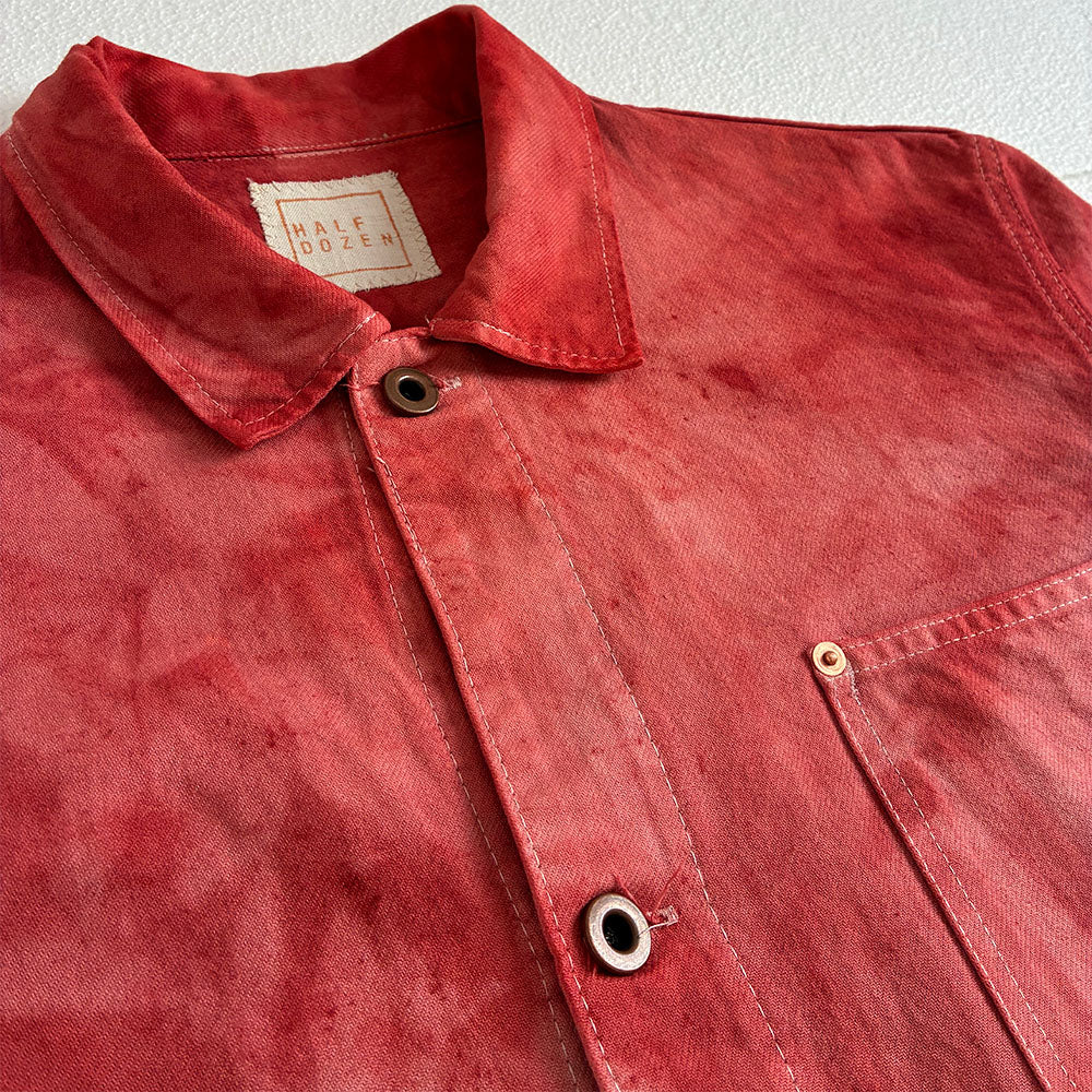 A close-up of a red washed cotton button-up shirt by Half Dozen, showing the collar, metal snap buttons, and a chest pocket against a light textured background.