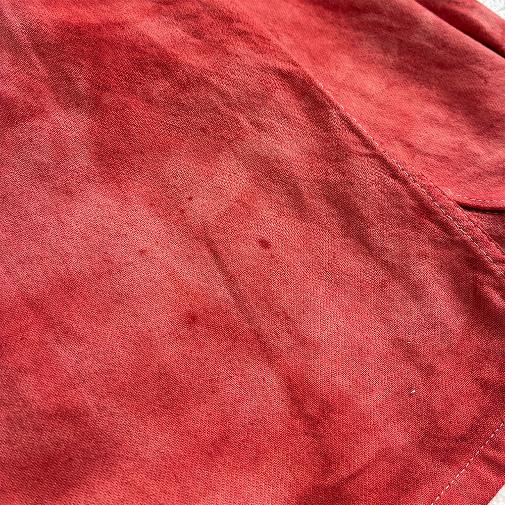 A close-up of a red chore coat fabric surface with visible texture, stitching, and a few small dark specks.