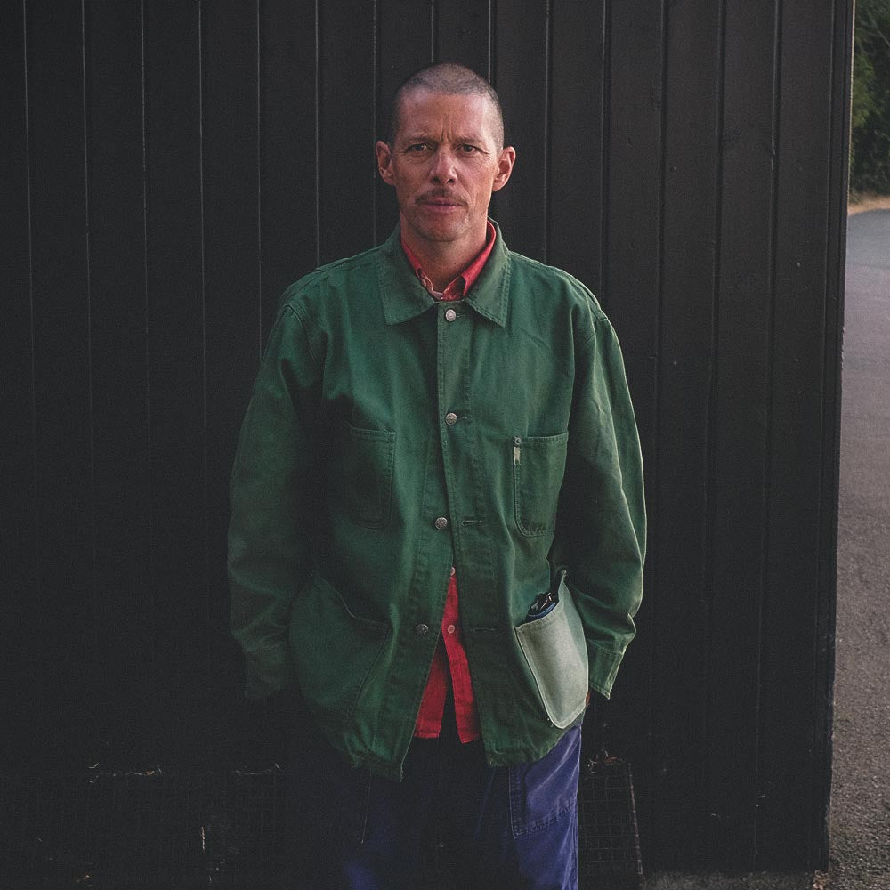 A man wearing a hand dyed green farmers utility jacket over a red shirt, blue trousers, and green rubber boots, standing in front of a dark wooden door.