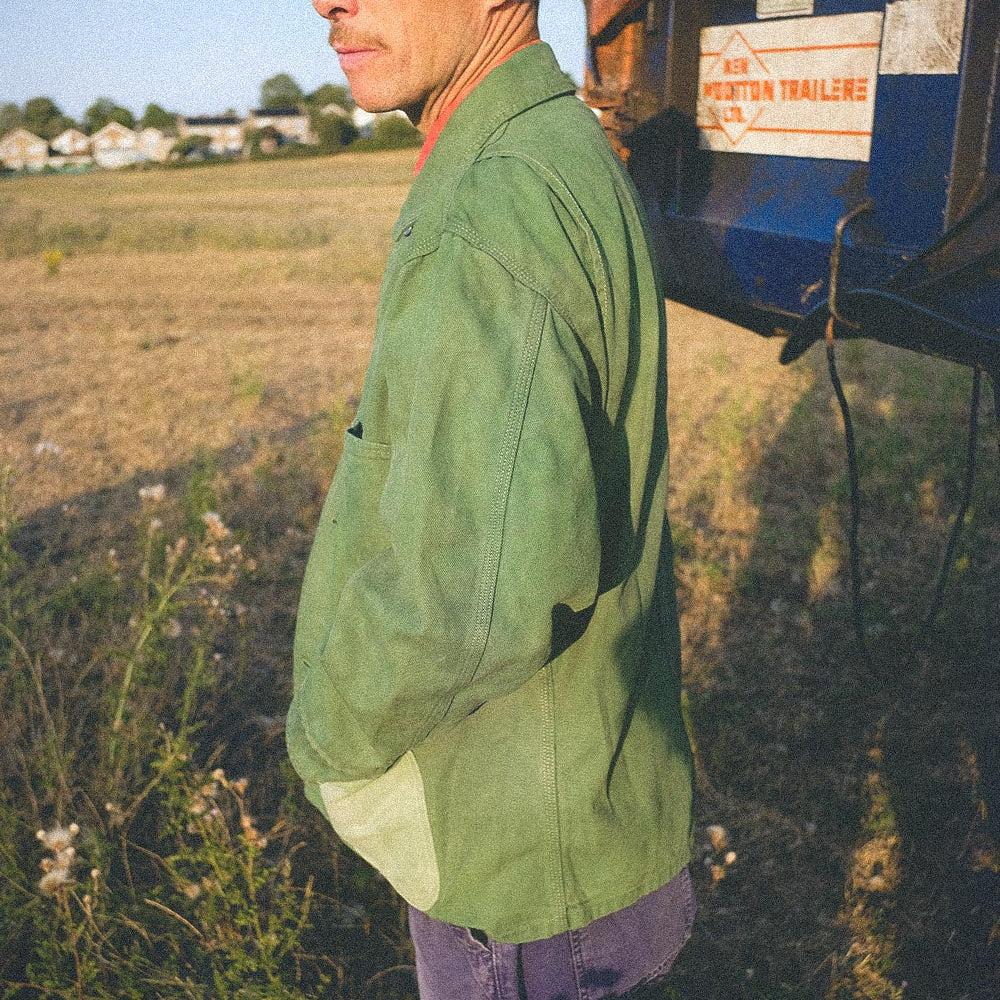 A man in a green work jacket and purple pants standing outdoors next to a blue trailer in a field.