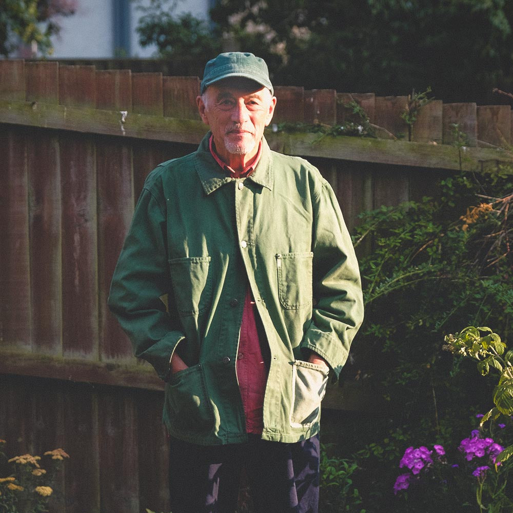 An older man standing outdoors in a garden or backyard, wearing a hand dyed green farmers jacket, dark trousers, black shoes, and a green cap. A wooden fence, plants, and potted flowers are visible behind him.