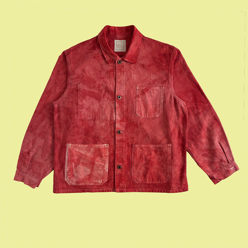 A red washed cotton chore jacket with four front pockets and a button-up front, copper donut buttons laid flat against a pale yellow background.