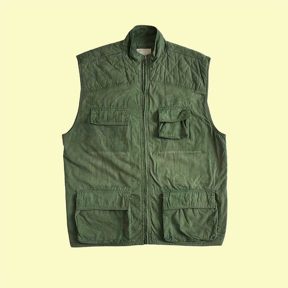 An olive green quilted utility gilet with a front zipper, a stand collar, and multiple flap pockets, shown flat against a pale yellow background.
