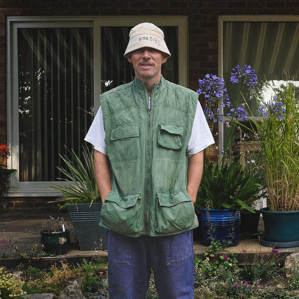 A man stands outdoors in front of a house, wearing a green gilet vest over a white t-shirt, blue jeans, and a beige bucket hat.