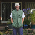 A man stands outdoors in front of a house, wearing a green gilet vest over a white t-shirt, blue jeans, and a beige bucket hat.