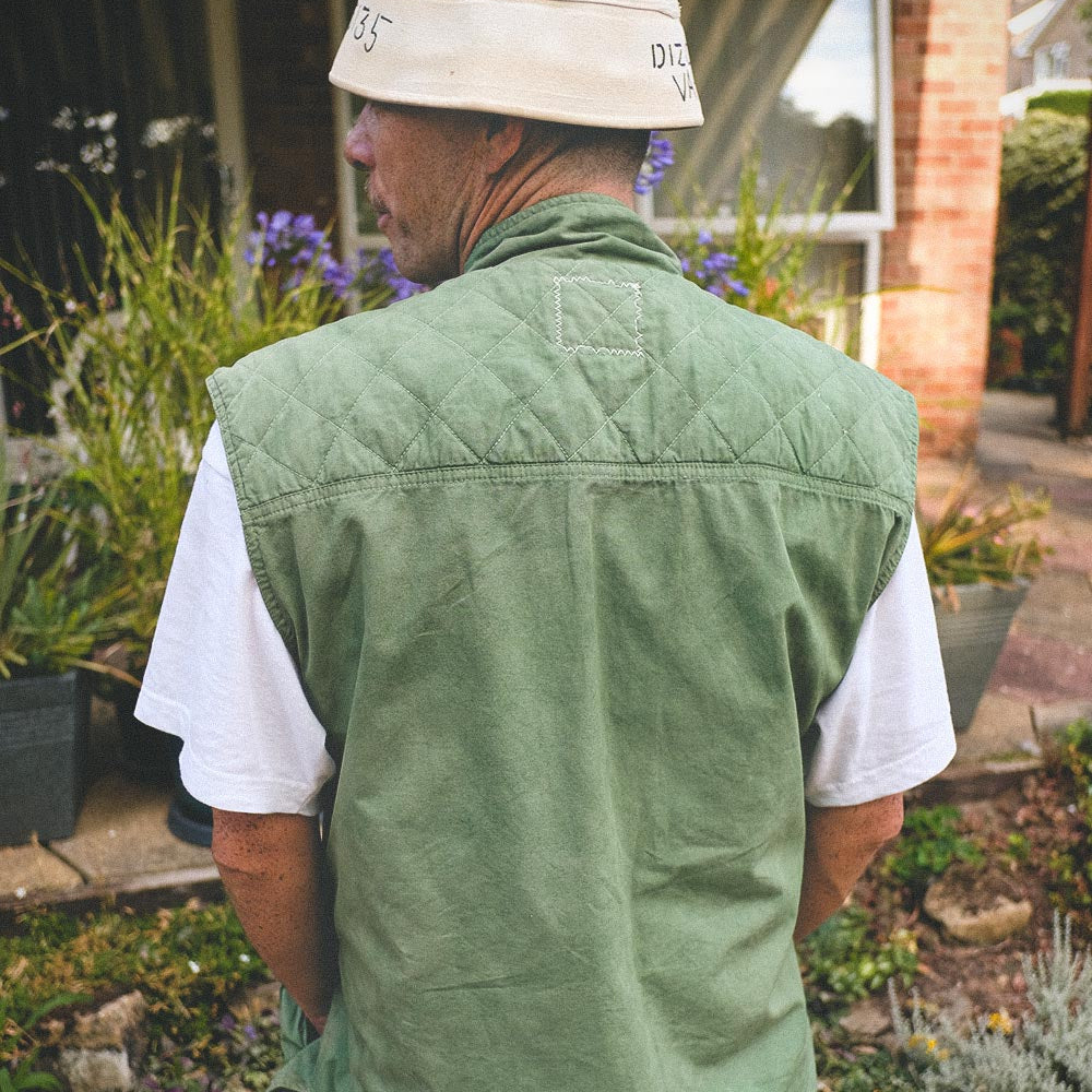 A man viewed from behind wearing a green quilted utility gilet over a white t-shirt, a beige bucket hat, and blue jeans, standing in a garden in front of a brick house.