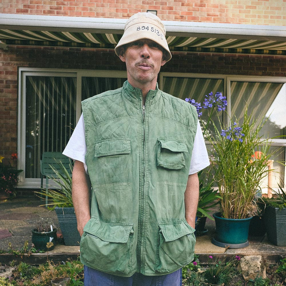 A man wearing a light green utility gilet over a white t-shirt and blue jeans, standing outdoors in front of a brick house with potted plants.