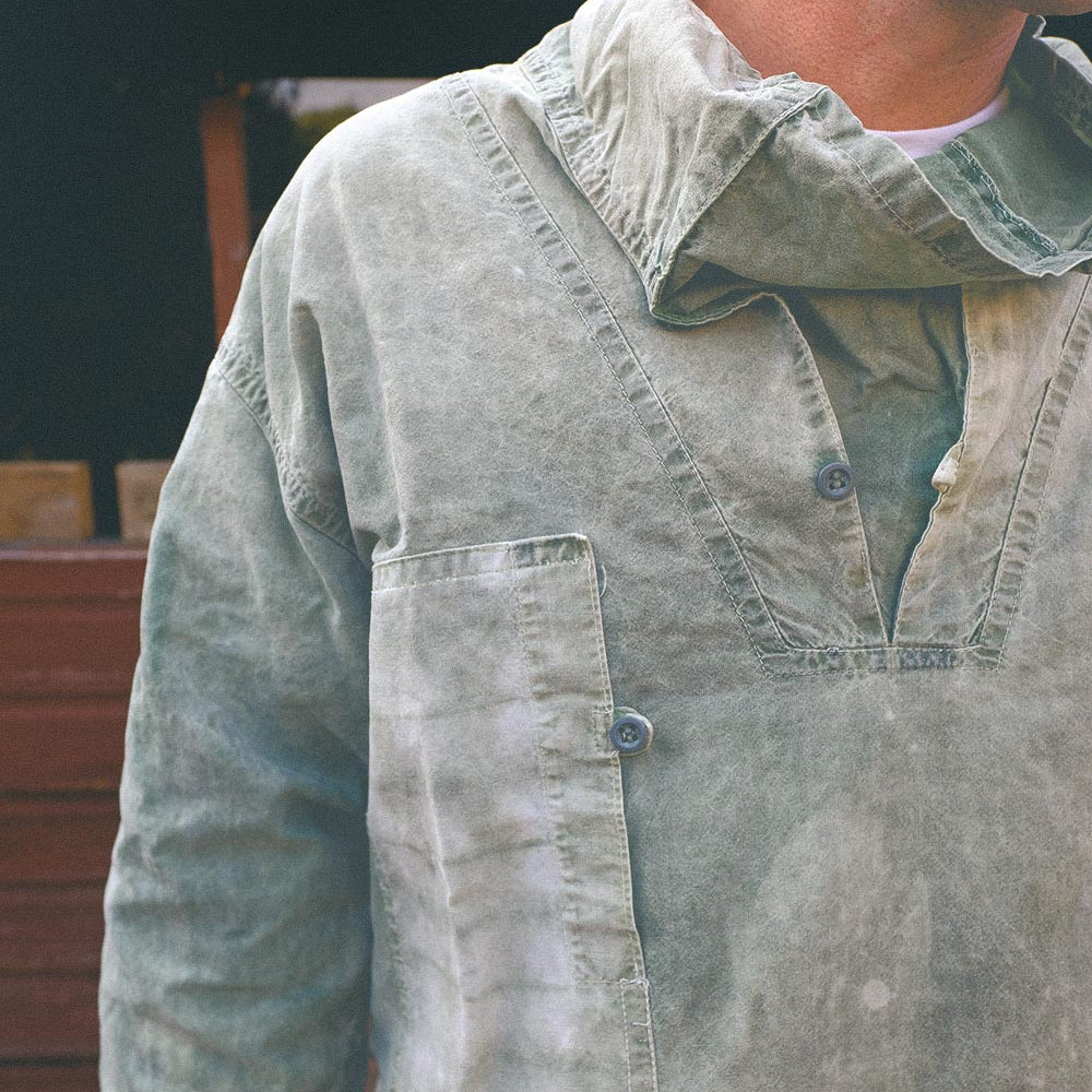 A close-up of a person wearing a faded green smock with a buttoned chest pocket, hand dyed and waxed, photographed outdoors.