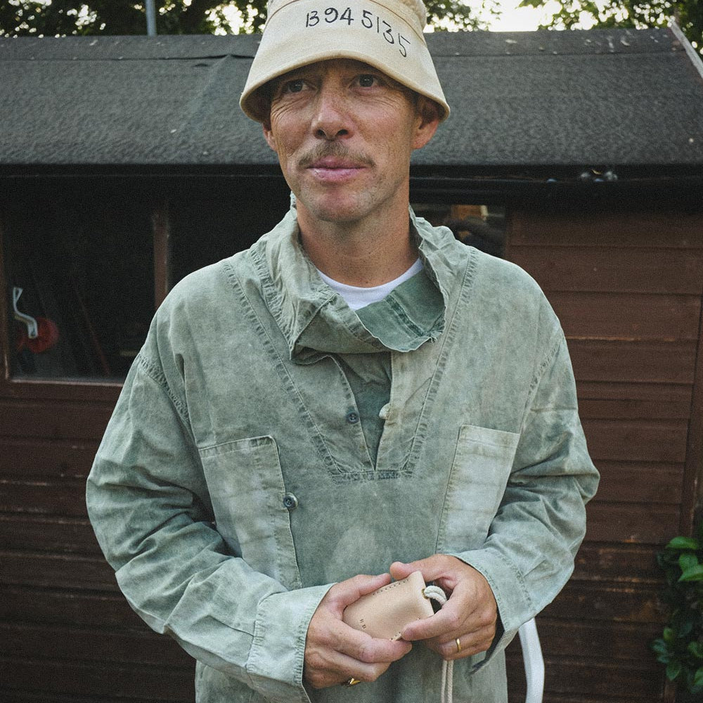 A man wearing a light beige bucket hat with black printed numbers and a hand dyed military replica smock, standing outdoors in front of a brown wooden shed.