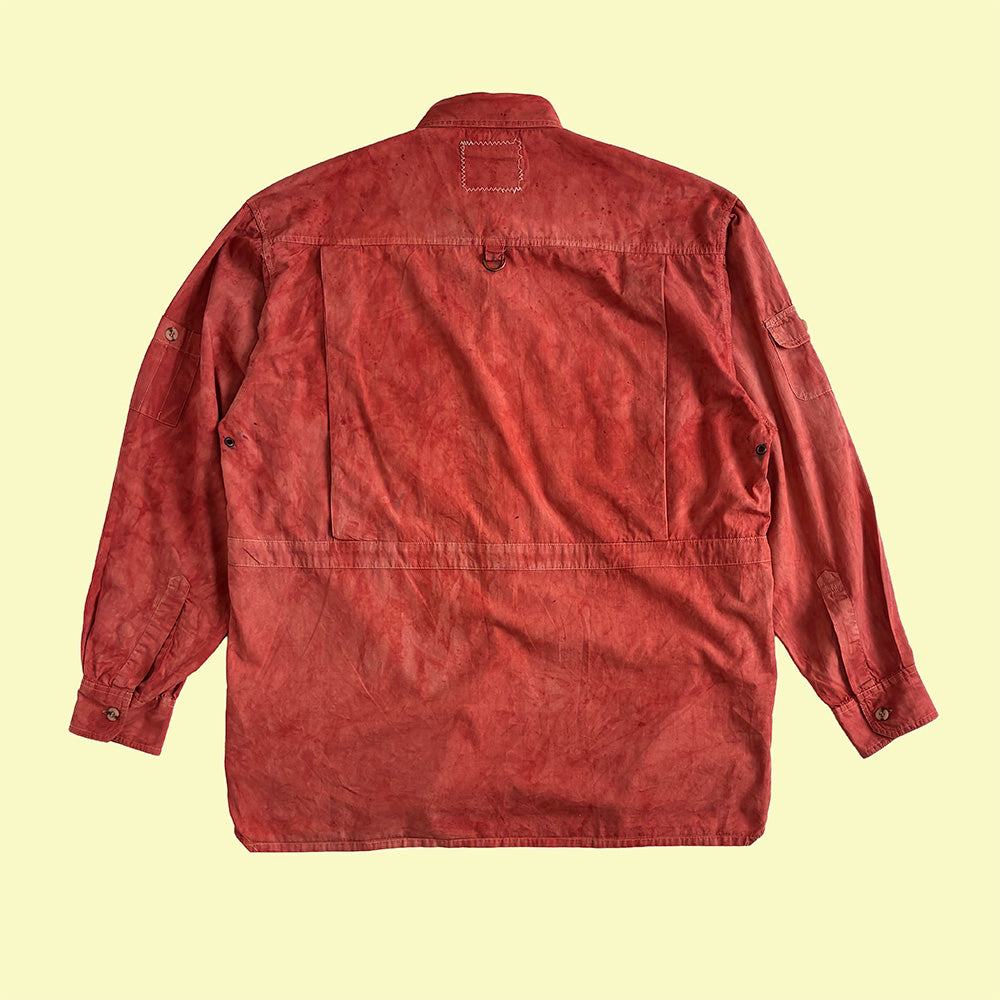 A red hand dyed cotton utility shirt shown from the back, laid flat against a pale yellow background.