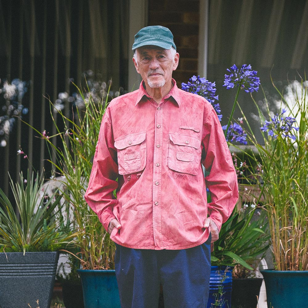 An older man standing outdoors in a garden or yard, wearing a coral pink long-sleeve button-up shirt with chest pockets, navy blue pants, a green cap, and dark shoes. Potted plants and a house exterior are visible in the background.