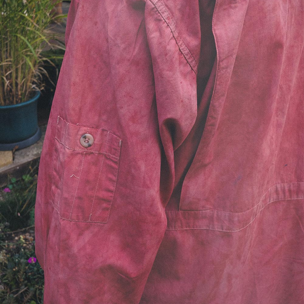 A close-up of a red and pink utility cotton overshirt jacket, showing the back pocket and shoulder area, photographed outdoors with garden plants in the background.
