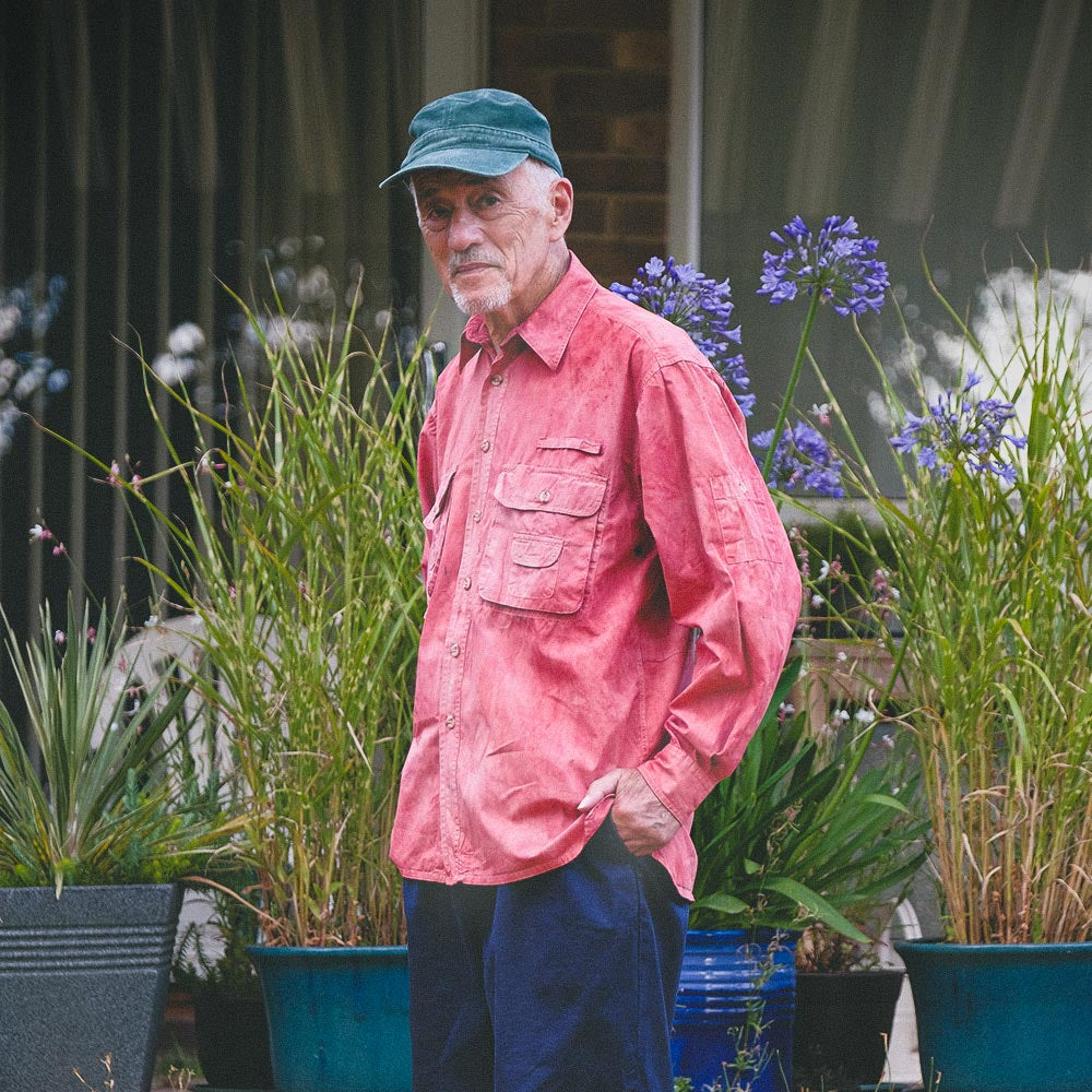An older man standing outdoors in a garden wearing a indian madder hand dyed pink and red long-sleeve button-up shirt, dark blue pants, a green cap, and black shoes.