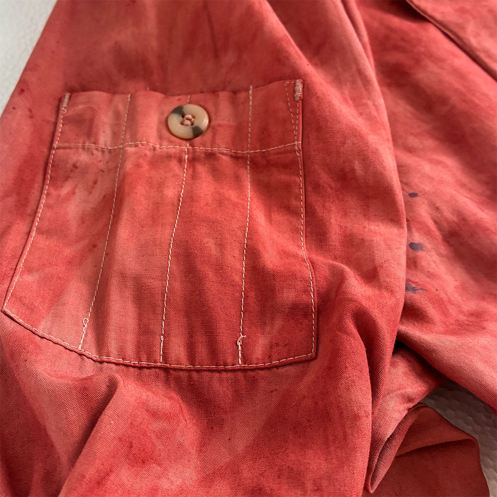 A close-up of a washed, hand dyed red-pink linen fabric garment, showing a buttoned chest pocket with vertical stitching.