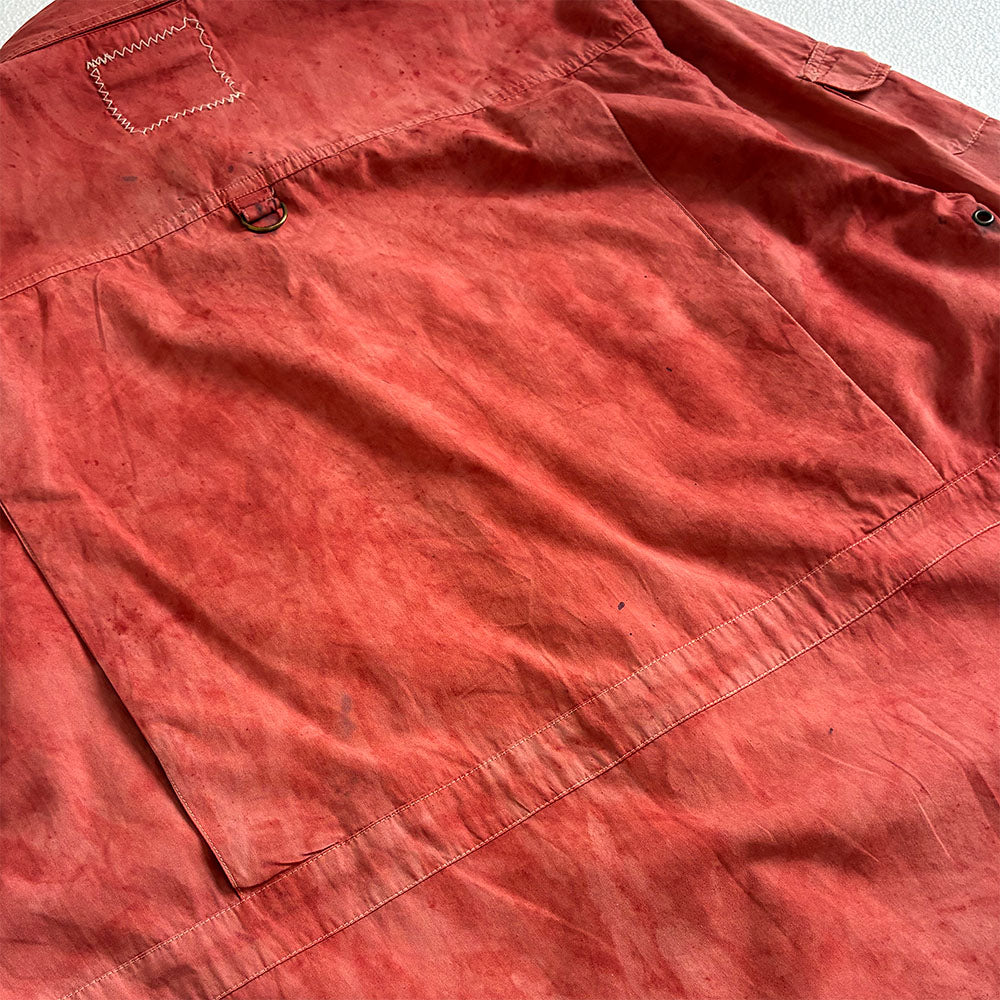 A close-up of a red-pink utility shirt laid flat, showing the fabric texture, a large front patch pocket, and a metal D-ring near the chest.