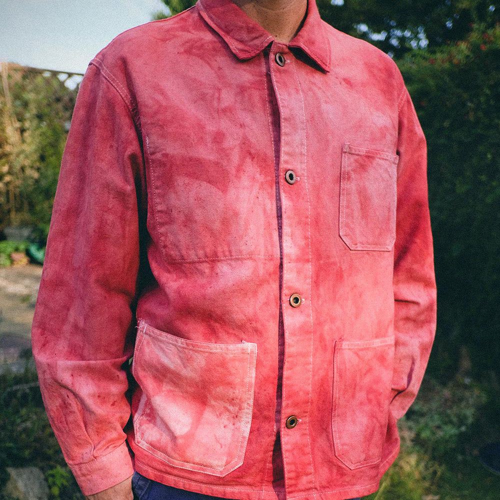 A person wearing a faded red-pink chore jacket with four front pockets, photographed outdoors against a blurred green garden background.