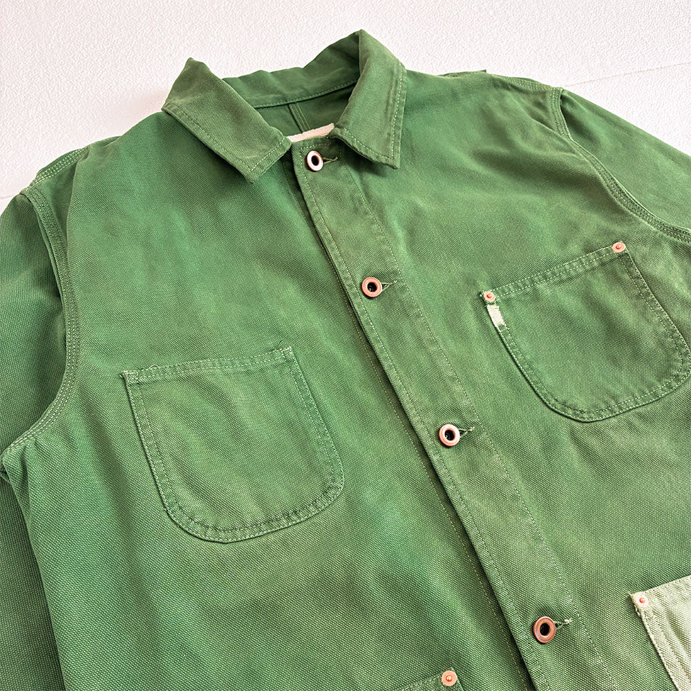 A green canvas weave button-up shirt jacket with two chest pockets and copper-colored donut buttons, laid flat on a white textured background.